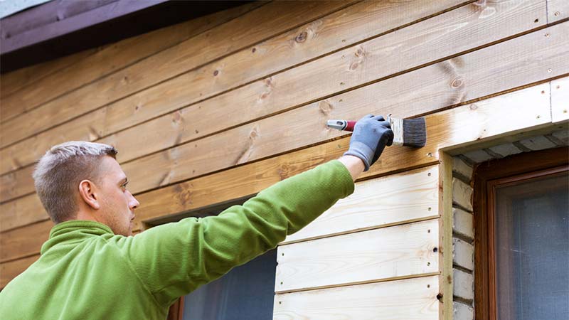 Man in groene trui brengt transparante beits aan op tuinhuis